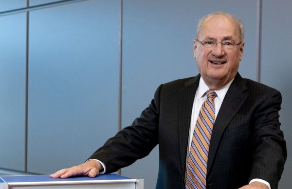 portrait-of-a-man-in-a-suit-smiling-at-camera-with-both-hands-on-top-of-gray-and-blue-rectangles.jpg