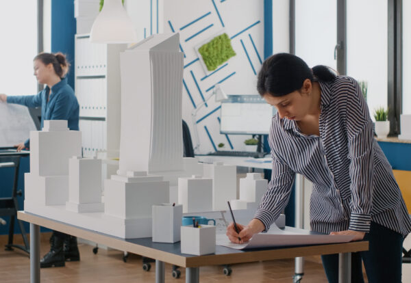 Woman architect analyzing blueprints plan on table to design building model and maquette. Engineer working on architectural development project with industrial construction tools.